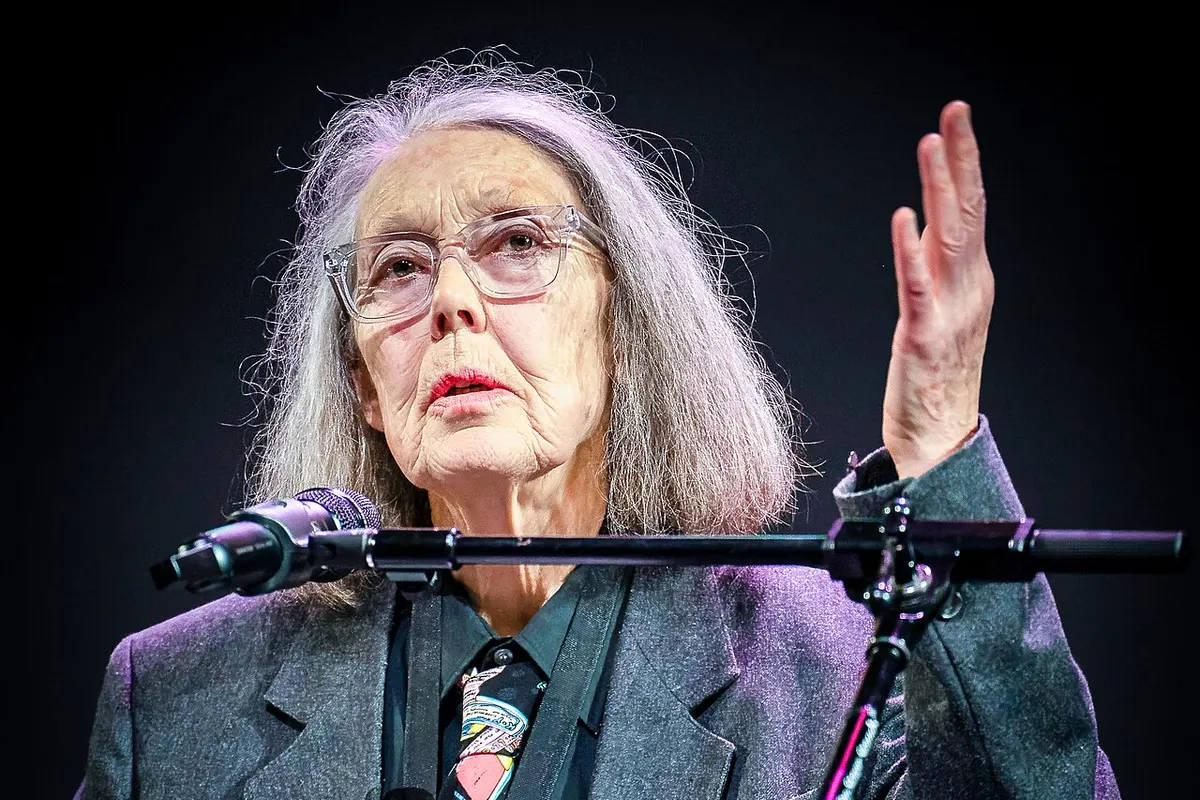 Anne Carson habla en un podio transparente con la mano derecha levantada